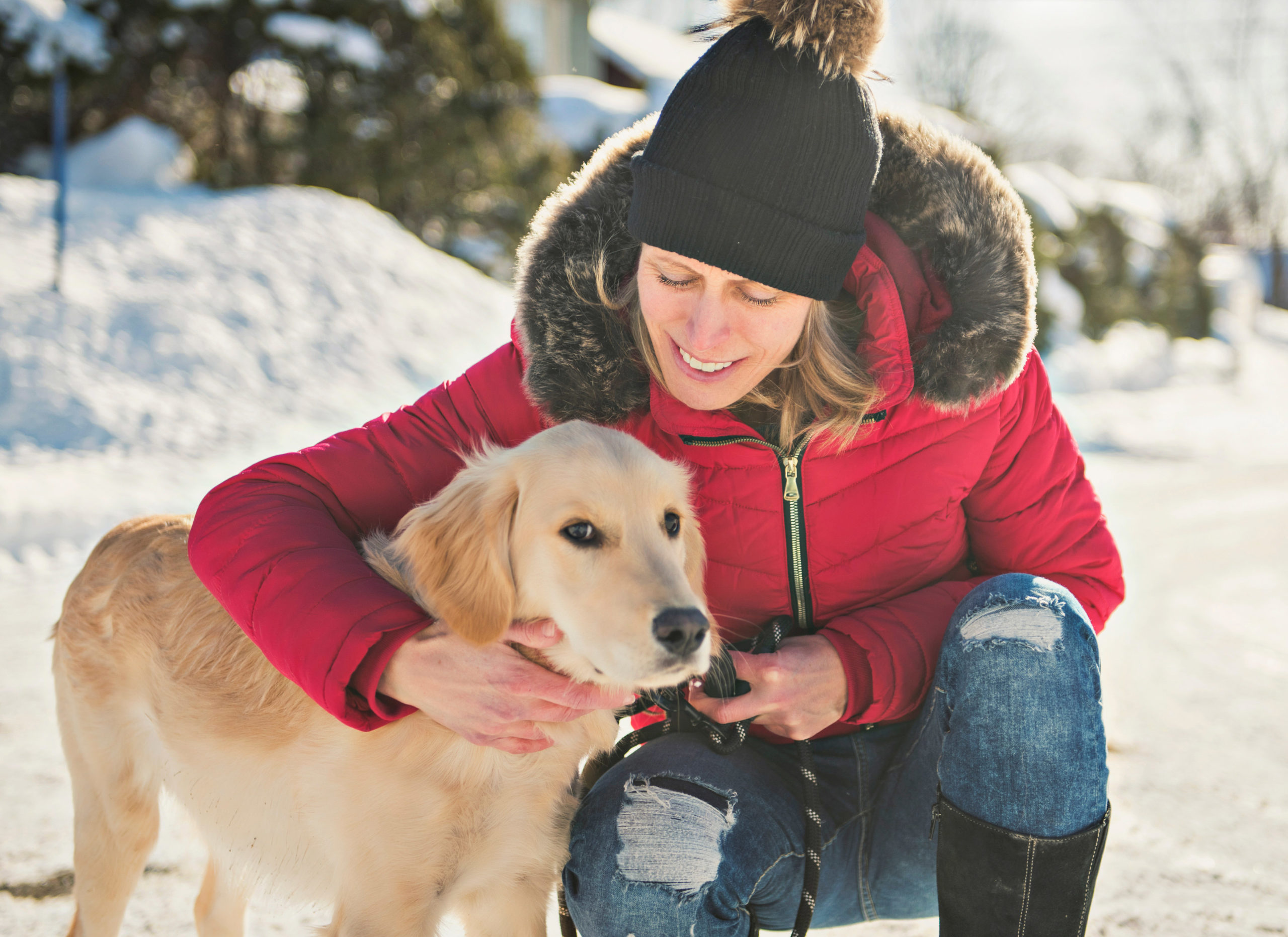 En voksen, blond dame med lue sitter på huk og klapper sin hund i snøen.